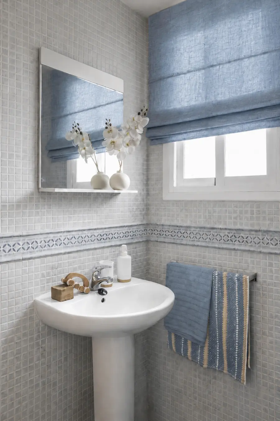 Baño con mosaico original y luz natural Baño con mosaico claro, lavabo de pedestal, espejo sin marco y estor azul sobre la ventana
